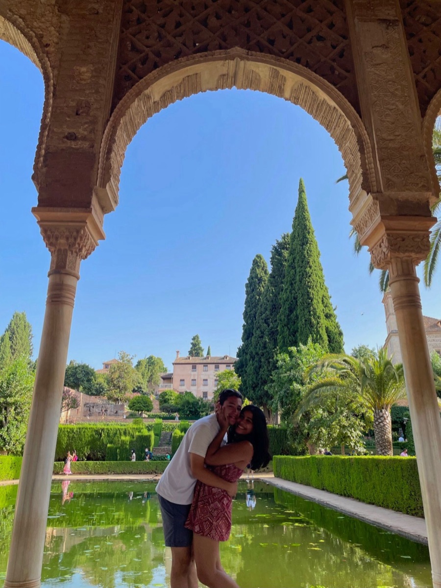 Marcos and Tanya hugging under stone arches by a garden pool